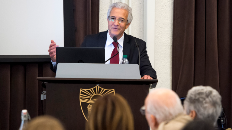 Picture of Omer Bartov, smiling behind a podium, left hand raised, gesturing.
