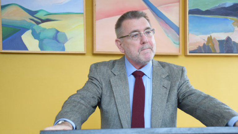 Picture of Peter Hayes lecturing. He has both hands on the podium. Behind him are three abstract pastel landscapes on a yellow wall.