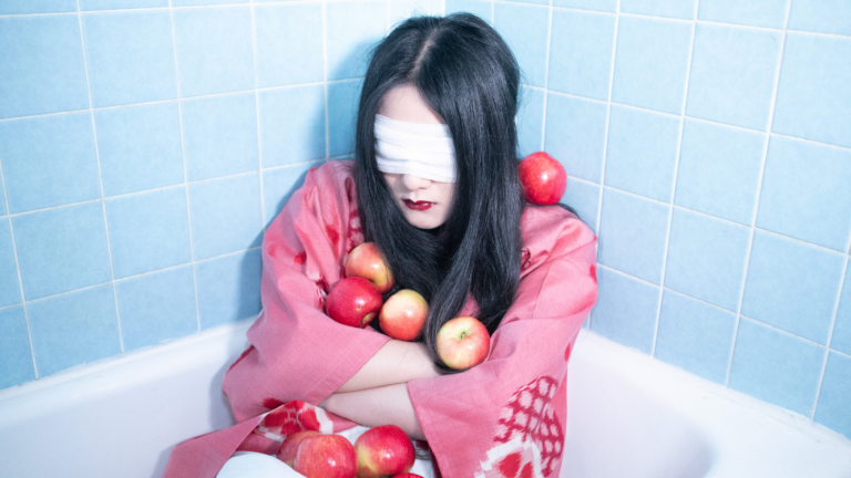 A blindfolded woman sits in a bathtub, covered in apples.
